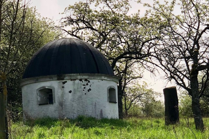 Small round building with dome in grassy area, surrounded by trees.